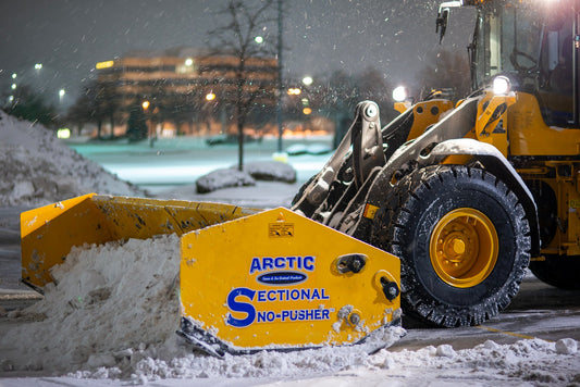 Snow plow with 'Arctic Sectional Snow Pusher' on a snowy street at night.