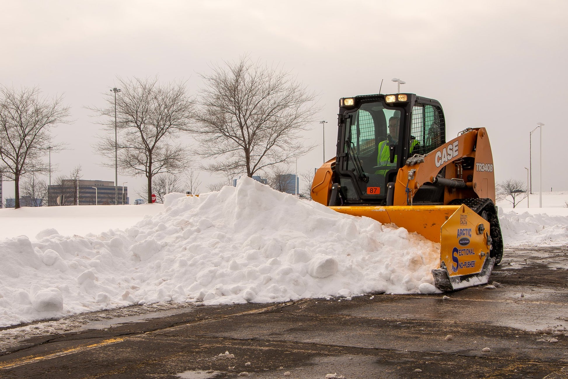 Snow plow clearing snow from a parking lot on a cloudy day.